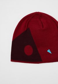 Gorro de punto rojo con un diseño geométrico que presenta un triángulo rojo oscuro y un detalle circular, acentuado con un triángulo azul claro.