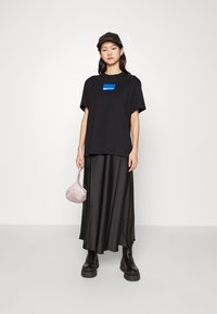 Camiseta negra de gran tamaño con gráfico azul, falda maxi de satén negra, botas negras gruesas y un pequeño bolso metálico. Diseño minimalista.