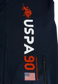Tessuto blu navy con testo bianco e arancione "USPA 90 Heritage Sportswear", logo giocatore di polo arancione e piccola bandiera americana vicino a una cerniera nera.