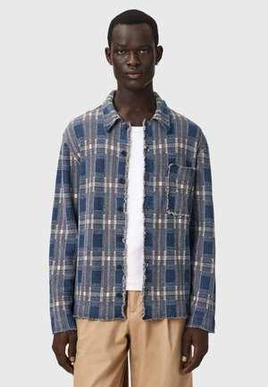 Mann trägt eine blau-beige karierte, geknöpfte Jacke mit ausgefransten Rändern, weißes T-Shirt und beige Hose, steht vor einem schlichten Hintergrund.