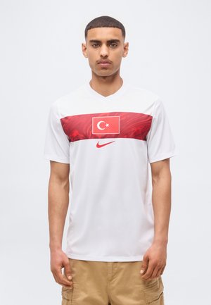 Giovane uomo che indossa una maglia sportiva bianca con una bandiera turca rossa e il logo Nike, abbinata a pantaloncini cargo marroni chiari.