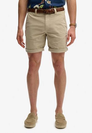 Man wearing rolled-up beige shorts, brown braided belt, blue floral shirt, watch, and beige loafers against white background.