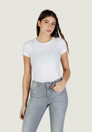 Donna che indossa una T-shirt aderente bianca di Armani Exchange e jeans grigio chiaro a vita alta, in piedi davanti a uno sfondo semplice.