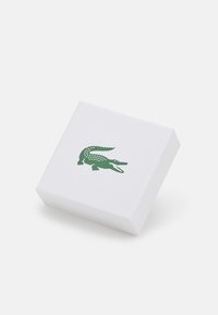 Hvid kasse med tekstureret overflade. Har et grønt krokodille-logo på fronten. Firkantet form, minimalistisk design.