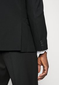 Zwarte colbert met een gestructureerde afwerking, voorzien van een enkele split, vier knopen op de mouwen en een wit manchet dat zichtbaar is.
