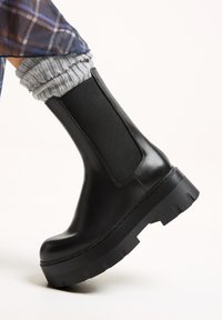 Gheată neagră din piele, cu panou elastic pe lateral, talpă groasă din cauciuc negru și șosete gri texturate care ies la vedere în partea de sus.