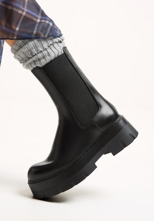 Fekete bőrbokacsizma elasztikus oldalsó betéttel, vastag fekete gumitalppal, és texturált szürke zoknival, amely a tetején kandikál ki.