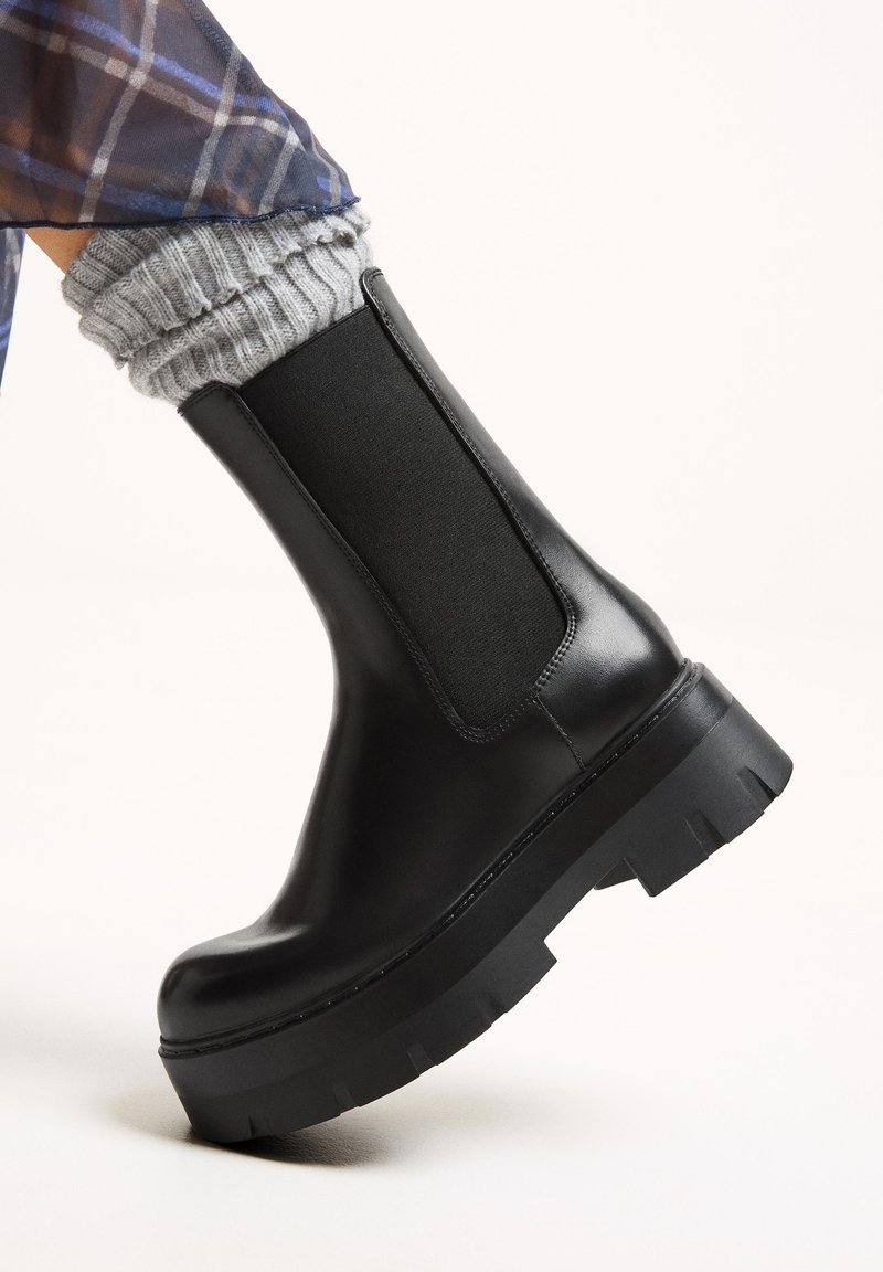 Gheată neagră din piele, cu panou elastic pe lateral, talpă groasă din cauciuc negru și șosete gri texturate care ies la vedere în partea de sus.
