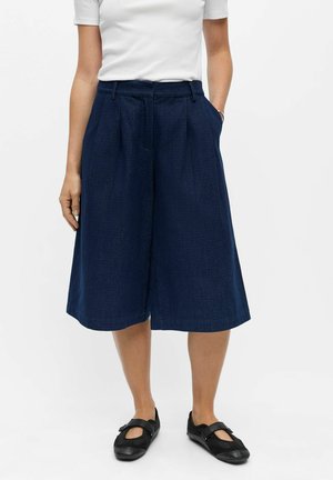 Personne portant un short large bleu marine jusqu’au genou, une chemise blanche à manches courtes et des chaussures noires de style Mary Jane.
