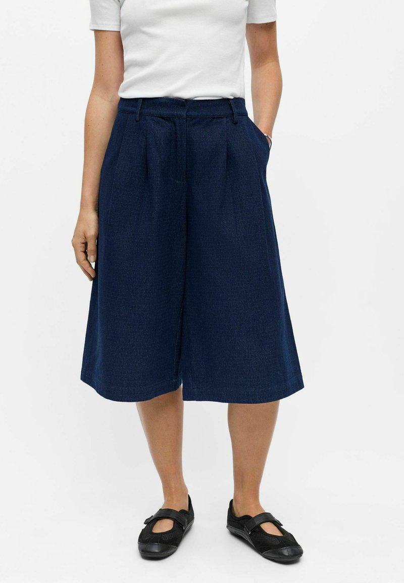 Personne portant un short large bleu marine jusqu’au genou, une chemise blanche à manches courtes et des chaussures noires de style Mary Jane.