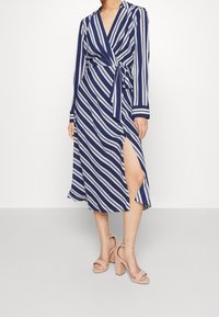 Pruhované zavinovací šaty v námořnické modré a bílé barvě s dlouhými rukávy, výstřihem do V, bočním rozparkem a odpovídajícím páskem. Modelka je doplnila sandály na vysokém podpatku ve světlé barvě.