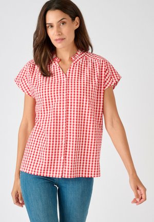 Mujer con blusa de cuadros vichy roja y blanca de manga corta con botones, cuello con volantes y vaqueros azules sobre un fondo liso.
