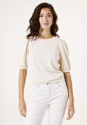 Haut à manches bouffantes de couleur crème, fabriqué dans un matériau doux, avec un décolleté arrondi et des poignets côtelés. Assorti à un jean blanc ajusté.