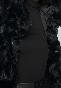 Sort faux fur jakke med tekstureret overflade, der har skinnende sort foring, knapper og en tætsiddende sort rullekrave bluse under.