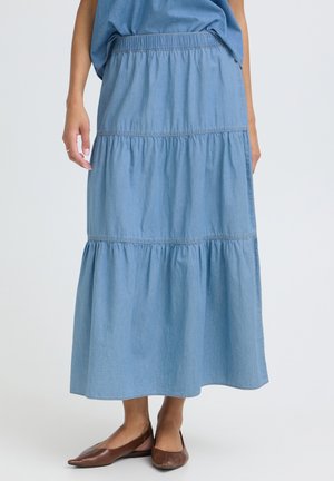 Kvinde iført en blå lagdelt midi-nederdel med elastisk talje og brune spidse flade sko, stående mod en ensfarvet baggrund.