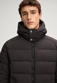 Giacca piumino nera con motivo a trapunta orizzontale, colletto alto, chiusura a zip e dettagli con bottoni a pressione. Tessuto morbido, design minimale.