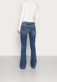 Personne portant un jean bootcut bleu et un haut blanc à manches longues côtelé, debout devant un fond uni gris clair.