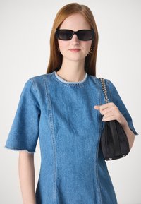 Denim-mekko, jossa on lyhyet hihat, revennyt kaulus ja pystysuorat saumat. Malli pitää kädessään pientä mustaa käsilaukkua, jossa on ketjuhihna. Tummat aurinkolasit.