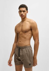 Brown patterned swim shorts made of lightweight fabric, featuring an elastic waistband with drawstring and a small logo patch on the side.