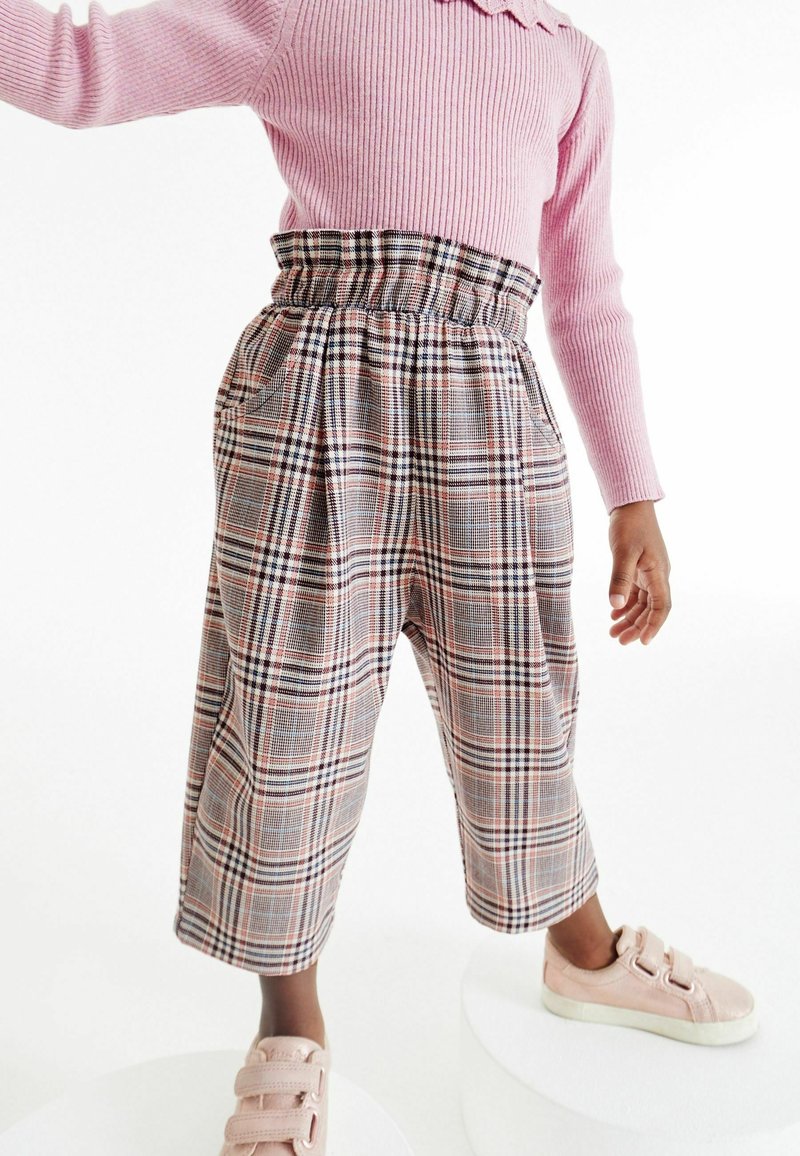 Enfant portant un pull côtelé rose, un pantalon à carreaux taille haute et des baskets rose clair, debout sur des plateformes blanches.