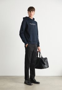 Mornarska hoodie s prednjim džepom i bijelim logom, uparen s crnim hlačama i crnom kožnom torbom. Model nosi crne cipele. Jednostavan, čist dizajn.