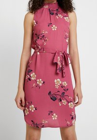 Růžové šaty bez rukávů s květinovým potiskem, s vysokým nařaseným výstřihem a opaskem v pase, nošené stojící osobou od ramen po kolena.