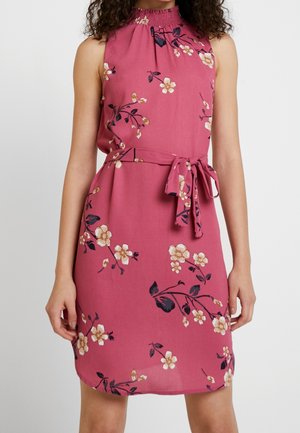 Hihaton vaaleanpunainen mekko, jossa on kukkakuosi, korkea kerätty kaula-aukko ja vyö, jota henkilö pitää yllään olkapäistä polviin saakka.