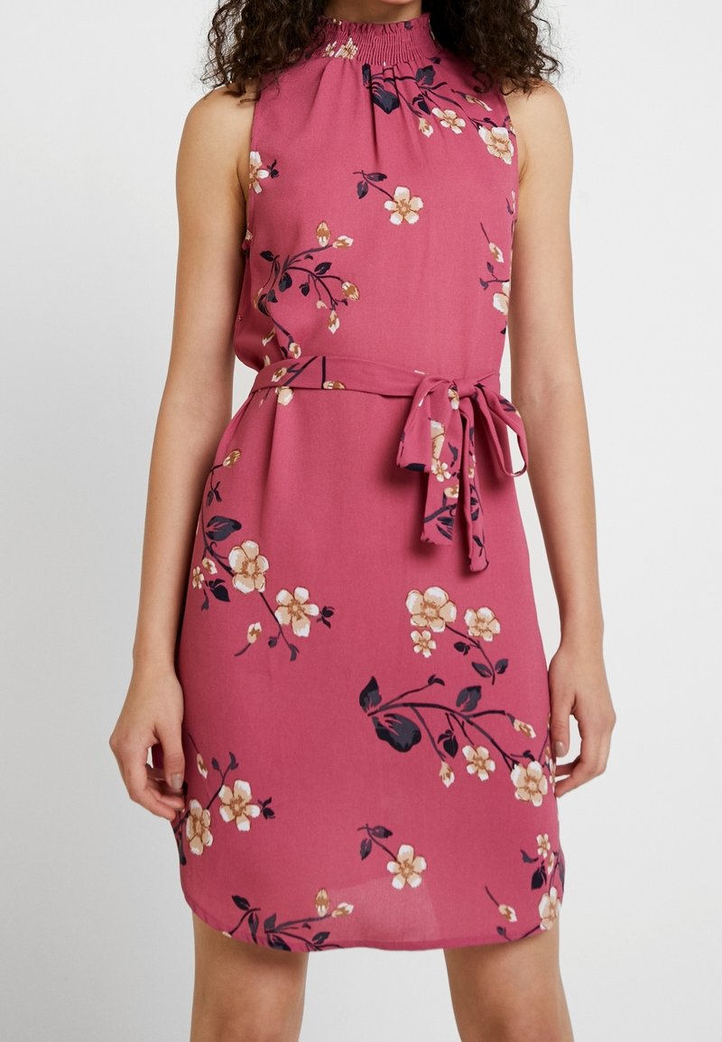 Růžové šaty bez rukávů s květinovým potiskem, s vysokým nařaseným výstřihem a opaskem v pase, nošené stojící osobou od ramen po kolena.