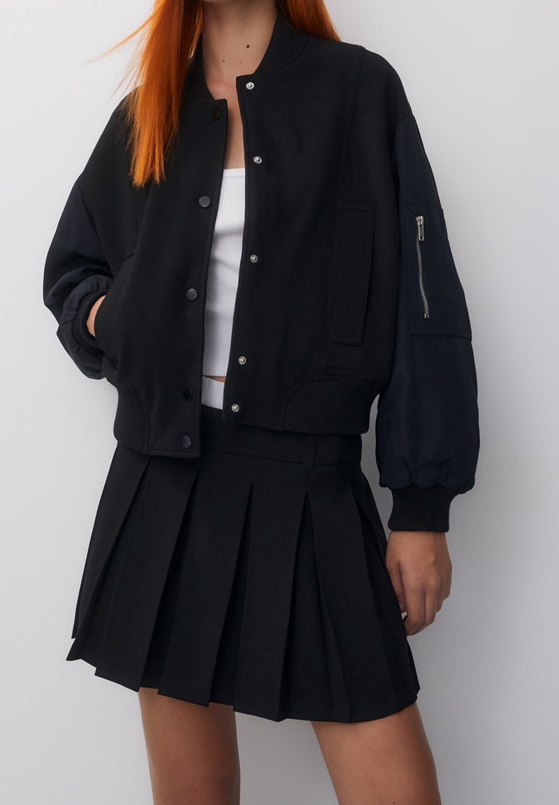 Blouson bomber noir avec boutons-pression, poignets côtelés et une poche zippée. Associé à une jupe noire plissée, les textures des deux tissus sont visibles.