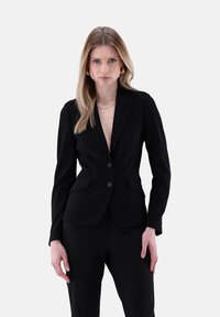 Schwarzes Blazer mit angepasstem Design, strukturiertem Stoff, zwei Knöpfen und spitzen Revers. Kombiniert mit maßgeschneiderten schwarzen Hosen für einen formellen Look.
