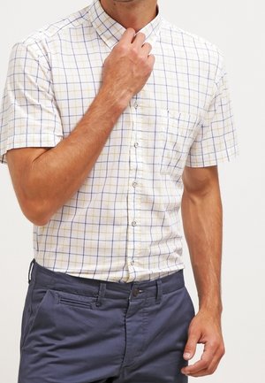 Homme ajustant son col, portant une chemise blanche à manches courtes avec un motif en grille jaune et bleue, et un pantalon bleu foncé, sur un fond uni.