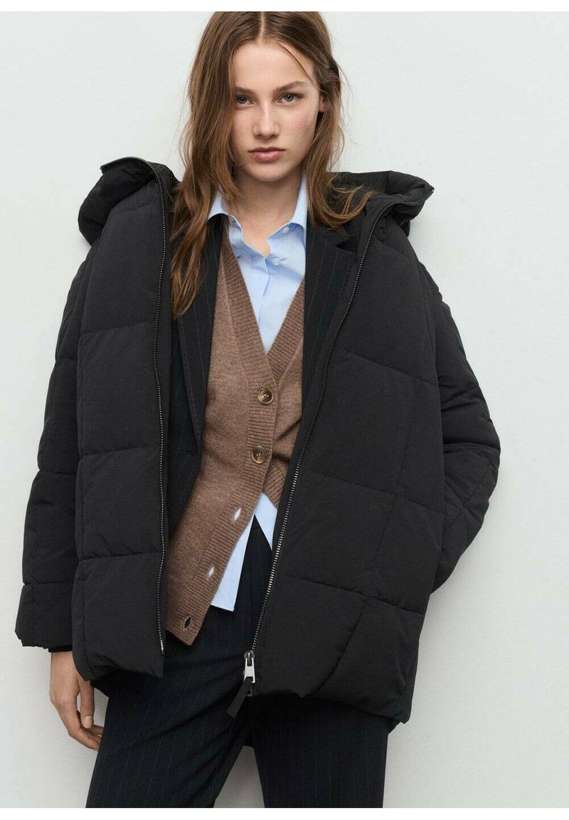 Chaqueta de plumas negra con un diseño acolchado y capucha, que presenta una cremallera delantera y bolsillos laterales, llevada sobre un cárdigan marrón y una camiseta azul.