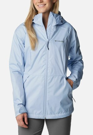 Donna che indossa una giacca antipioggia Columbia azzurro chiaro con cappuccio, zip frontale e mani nelle tasche, sorridendo leggermente.