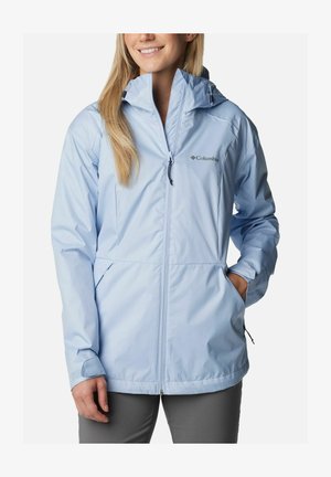 Donna che indossa una giacca antipioggia Columbia azzurro chiaro con cappuccio, zip frontale e mani nelle tasche, sorridendo leggermente.
