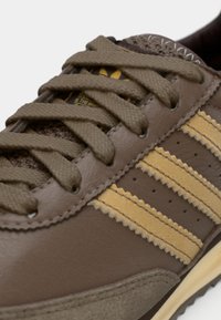 Smeđa kožna tenisica s teksturiranim detaljima, s tri žute pruge, okruglim vezicama i gumenim potplatom. Prisutne su suptilne perforacije.