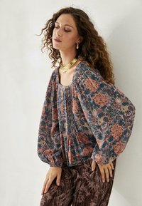 Blusa de tejido oscuro con patrón floral, con pliegues y mangas abullonadas. Combinada con pantalones marrones texturizados. Lleva un collar llamativo de oro.
