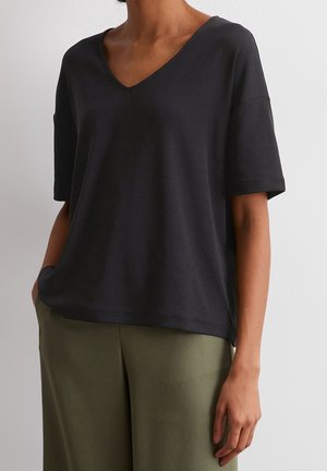 Kobieta w luźnej czarnej bluzce z krótkim rękawem i dekoltem w serek oraz oliwkowych spodniach, stojąca z jedną ręką w kieszeni na białym tle.