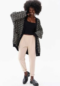 Zwarte cardigan met een wit geometrisch patroon, beigekleurige broek en zwarte instappers. Gelaagde gebreide stof, oversized pasvorm en opgerolde mouwen.