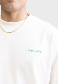 T-shirt en coton blanc à col rond, avec le texte brodé en vert "Redefined Rebel" sur la poitrine et une coupe décontractée.