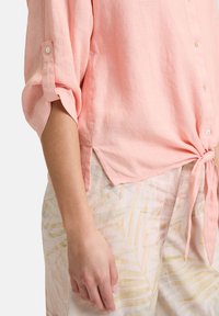 Lys pink linned skjorte med rullede ærmer, knaplukning og bundet for-ends. Sammen med mønstrede, lyse bukser med palmeblade.