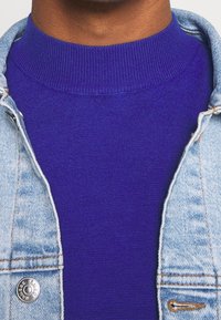 Lila stickad tröja med en hög ribbad krage, lager under en ljusblå jeansjacka. Jackan har en detalj med metallknapp.