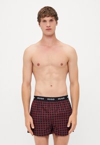 Shorts de boxeur noirs avec un motif à carreaux rouges, dotés d'une ceinture noire avec un logo "HUGO" blanc et un détail de bouton à l'avant.