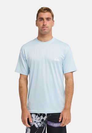 Hombre con camisa de manga corta azul claro y pantalones cortos negros con patrón y logo morado de Quiksilver, de pie frente a un fondo liso.