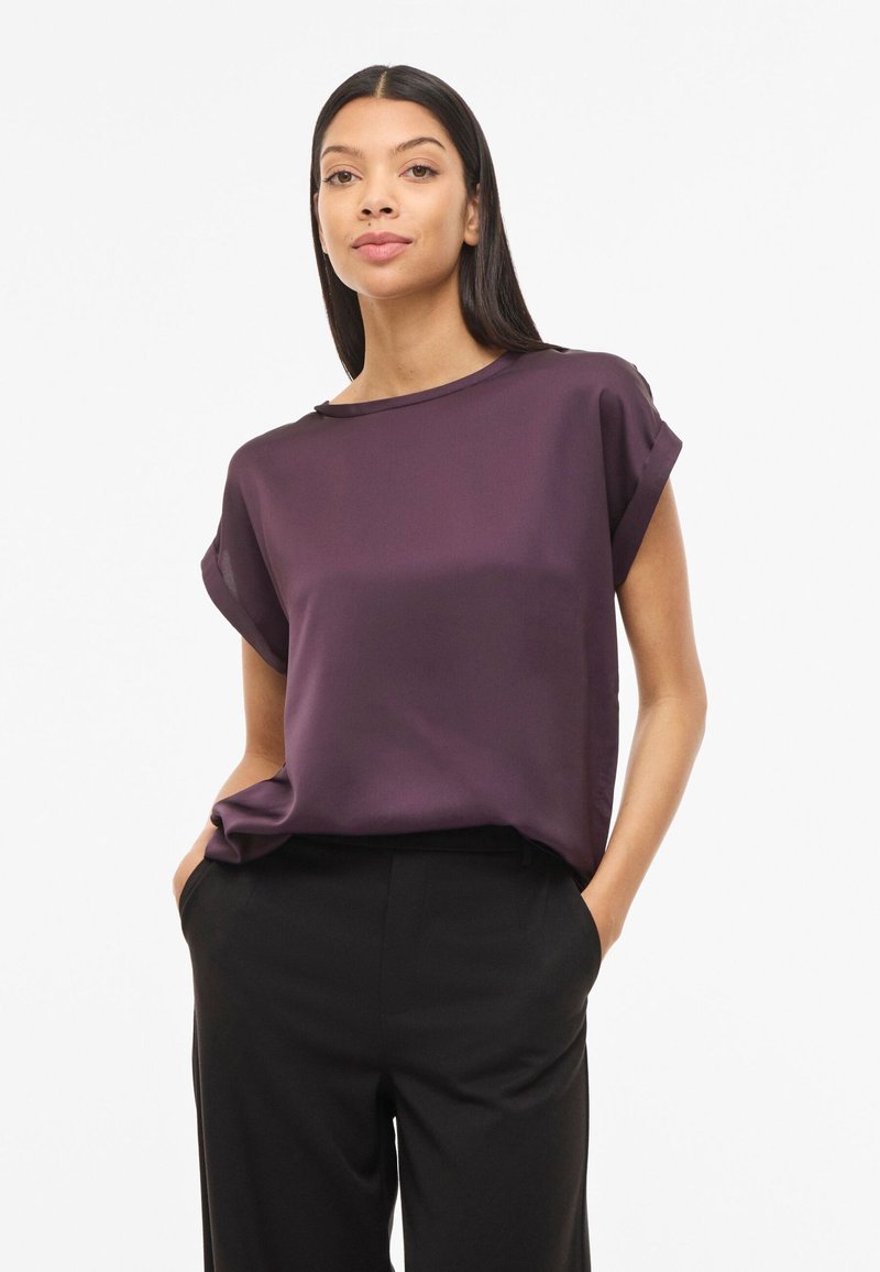 Kvinde med langt mørkt hår iført en løs, lilla bluse med korte ærmer og sorte bukser, hænderne i lommerne, neutral ansigtsudtryk.