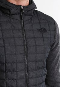 Mies, joka on pukeutunut mustaan, vetoketjulliseen, vuorattuun The North Face -takkiin hupulla, joka paljastaa ylärinnan ja kaulan alueen.