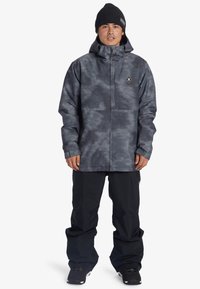 Šedá vzorovaná nepromokavá bunda s vysokým límcem, předním zipem a logem, v kombinaci s černými sněhovými kalhotami a botami.