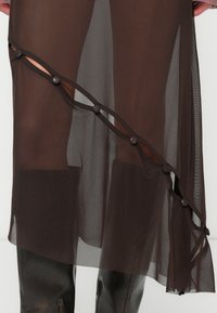 Barna áttetsző anyagból készült ruha réteges kialakítással, fekete kerek gombokkal az oldalon és folydogáló textúrával.