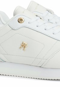 Zapatilla de cuero blanca con un emblema dorado, ojales redondos, paneles laterales texturizados y una suela blanca contrasta con diseño de zigzag.