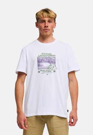 Homme blond portant un t-shirt blanc Billabong avec un motif de surf violet et vert et un short kaki, debout devant un fond uni.