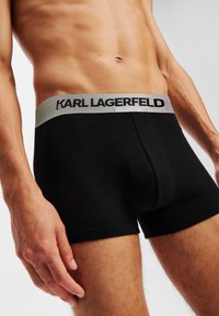 Crne bokserice z sivim elastičnim pasom, na katerem je napisan "KARL LAGERFELD." Narejene so iz gladke mešanice bombaža in imajo prilegajoč dizajn.